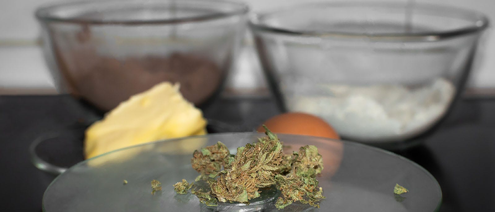 Cannabis flower on a scale in front of a bowl of flower, cocoa powder, butter, and an egg